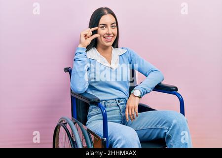 Belle femme aux yeux bleus assis sur un fauteuil roulant souriant et confiant gestante avec la main faisant un petit signe de taille avec les doigts regardant et le venu Banque D'Images