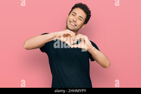 Jeune homme beau portant décontracté t-shirt noir souriant dans l'amour faisant coeur symbole de forme avec les mains. Concept romantique. Banque D'Images