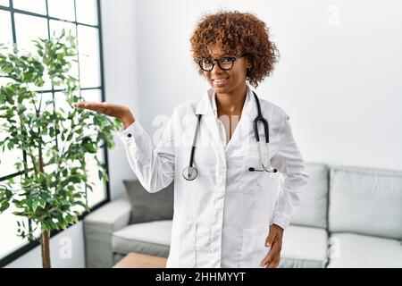 Jeune femme afro-américaine portant l'uniforme du médecin et le stéthoscope souriant gai présentant et pointant avec la paume de la main regardant l'appareil photo. Banque D'Images