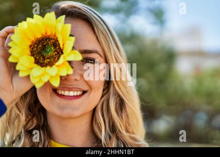 Jeune femme blonde souriant avec du tournesol à l'œil au parc Banque D'Images