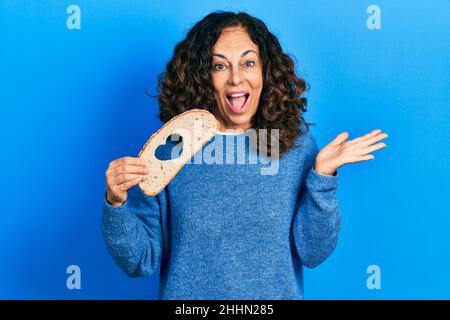 Femme hispanique d'âge moyen tenant le pain de pain avec la forme de coeur célébrant la réalisation avec le sourire heureux et l'expression de gagnant avec la main levée Banque D'Images