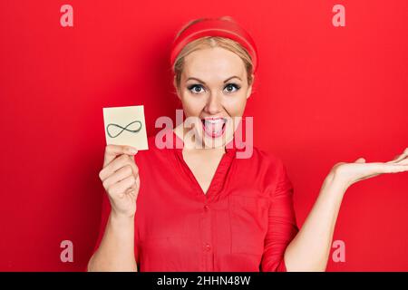 Jeune femme blonde tenant un symbole infini de rappel célébrant la victoire avec le sourire heureux et l'expression du gagnant avec les mains levées Banque D'Images