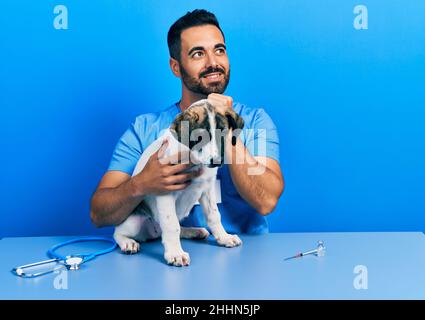 Beau vétérinaire hispanique avec la barbe en train de vérifier la santé du chien avec la main sur le menton en pensant à la question, l'expression pensive. Souriant et réfléchi Banque D'Images