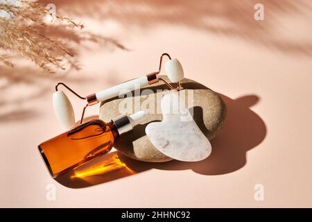 Massage facial au quartz blanc rouleau de Jade, Gua Sha et huile essentielle sur une pierre de galet de rivière sur un fond beige.CONCEPT DE soins de beauté SPA Banque D'Images