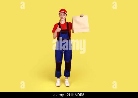 Portrait complet d'une femme de messagerie tenant le colis, livrant la nourriture dans un sac en papier, services de courrier postal, portant une combinaison et une casquette.Studio d'intérieur isolé sur fond jaune. Banque D'Images