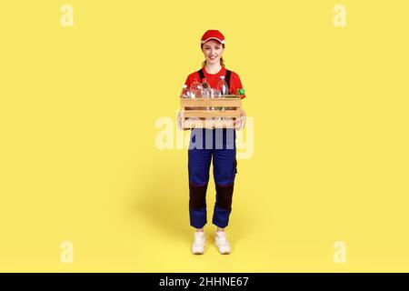 Portrait complet de la femme positive satisfaite volontaire debout avec une boîte pleine de bouteilles en plastique vides, portant une combinaison et un capuchon rouge.Studio d'intérieur isolé sur fond jaune. Banque D'Images