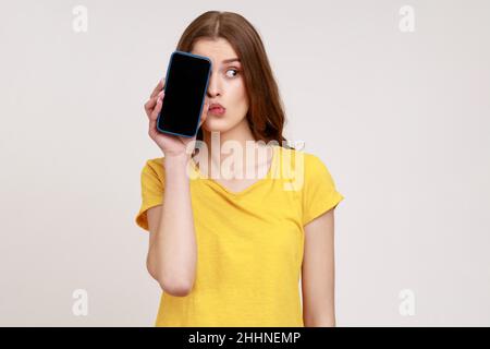 Regardez, annoncez ici.Portrait de drôle de femme de jeune âge en T-shirt couvrant la moitié du téléphone avec écran blanc, a l'expression de doute réfléchie.Prise de vue en studio isolée sur fond gris. Banque D'Images