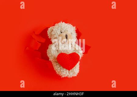 Ours en peluche tenant le coeur rouge Hug dans les pattes.L'ours en peluche sort des trous de papier déchirés avec le symbole coeur d'amour pour la Saint-Valentin isolé sur le rouge Banque D'Images