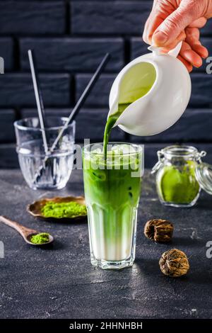 le barista verse le thé matcha dans un verre avec du latte matcha mélangé avec des glaçons avec un mur de brique à l'arrière-plan et du thé matcha en poudre dans du verre Banque D'Images