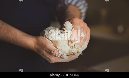 Un homme dans un tablier bleu pétrit la pâte à pain avec ses mains.Cuisine maison.Cuisson de la pâte dans la cuisine. Banque D'Images