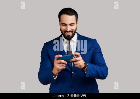 Ravi barbu homme utilisant le téléphone portable avec une expression positive heureuse, jouant à un jeu vidéo sur le téléphone portable, portant le costume de style officiel.Prise de vue en studio isolée sur fond gris. Banque D'Images