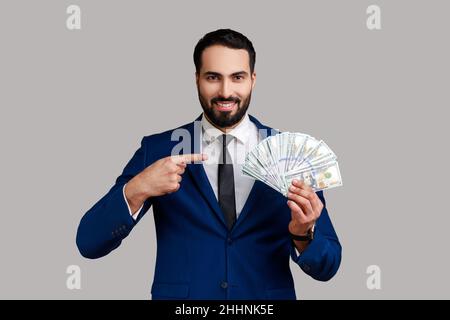 Riche barbu homme pointant sur des billets de banque en dollars et regardant avec une expression heureuse, prêt bancaire, épargne financière, portant un costume de style officiel.Prise de vue en studio isolée sur fond gris. Banque D'Images