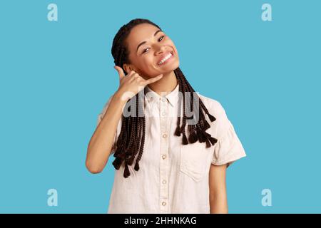 Jeune femme positive avec des dreadlocks noirs montrant le geste de téléphone, appelez-moi signe, demande le numéro de téléphone, se sent heureux, portant une chemise blanche.Studio d'intérieur isolé sur fond bleu. Banque D'Images