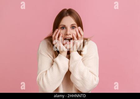 Portrait de femme blonde positive regardant la caméra de nat avec excitation, gardant les mains sur les joues, agréable surprise, portant le chandail blanc.Studio d'intérieur isolé sur fond rose. Banque D'Images