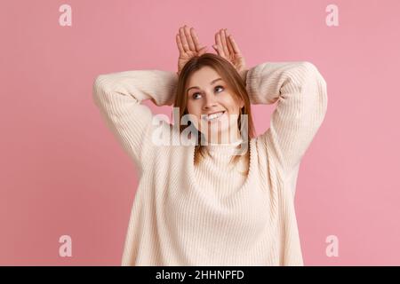 Portrait d'une femme blonde faisant des oreilles de lapin drôles avec les mains sur la tête, comportement puéril, humeur optimiste ludique, portant un chandail blanc.Studio d'intérieur isolé sur fond rose. Banque D'Images