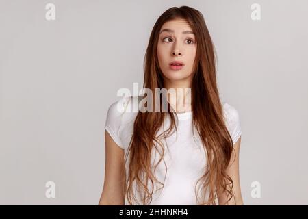 Drôle de jeune femme avec des cheveux foncés faisant le visage comique stupide avec les yeux croisés, pensant intensément à l'aspect stupide et confus, portant T-shirt blanc.Prise de vue en studio isolée sur fond gris. Banque D'Images