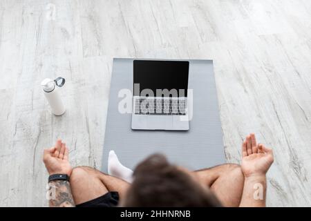 Jeune caucasien mimite dans la position de lotus est assis sur le sol sur le tapis, regarde PC avec écran vide Banque D'Images