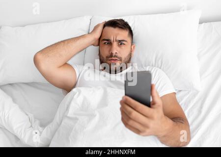 L'homme musclé européen millénaire défait se trouve sur un lit blanc, tient la tête à la main et lit l'actualité sur un smartphone Banque D'Images