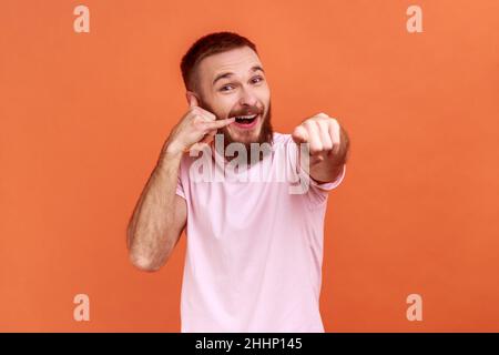 Portrait de l'homme barbu tenant les doigts près de l'oreille montrant le geste du téléphone regardant et pointant vers l'appareil photo, attendant votre appel, portant un T-shirt rose.Studio d'intérieur isolé sur fond orange Banque D'Images