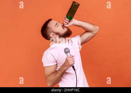 Portrait d'un homme barbu qui passe du temps au karaoké, tient un microphone en main, boit une boisson alcoolisée, porte un T-shirt rose.Studio d'intérieur isolé sur fond orange. Banque D'Images