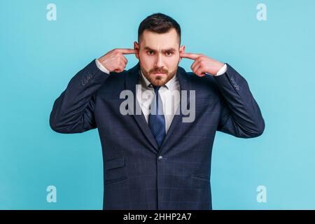 Je ne veux pas ici.Portrait d'homme d'affaires portant un costume officiel debout et tenant les doigts sur son oreille, sons désagréables.Studio d'intérieur isolé sur fond bleu. Banque D'Images