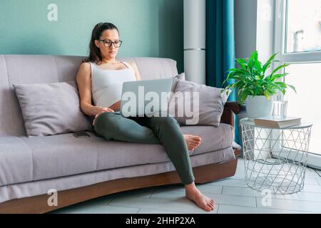 Jeune femme enceinte concentrée utilisant un ordinateur portable tout en étant assise sur un canapé à la maison Banque D'Images