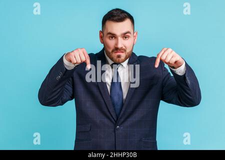 Venez-moi ici maintenant.Jeune adulte barbu homme en costume pointant les doigts vers le bas, exigeant approche à lui immédiatement, ayant le contrôle de la situation.Studio d'intérieur isolé sur fond bleu. Banque D'Images