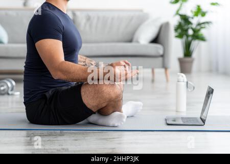 Homme musclé européen millénaire méditant en position lotus, assis sur un tapis avec ordinateur portable dans le salon Banque D'Images