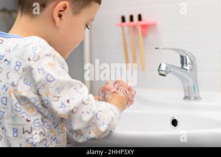 Un garçon de sept ans en pyjama se lave les mains à la maison dans une salle de bains lumineuse au-dessus d'un lavabo sur fond de carreaux et d'un robinet.Sélection Banque D'Images