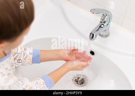 Un garçon de sept ans en pyjama se lave les mains à la maison dans une salle de bains lumineuse au-dessus d'un lavabo sur fond de carreaux et d'un robinet.Sélection Banque D'Images