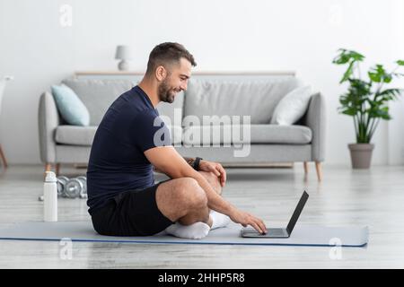 Joyeux jeune caucasien muscun gars assis dans la position de lotus sur le tapis et regarde l'ordinateur portable Banque D'Images