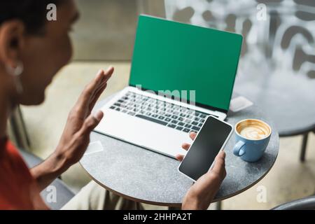Un travailleur indépendant afro-américain flou ayant un appel vidéo sur smartphone près d'un ordinateur portable et un café dans un café Banque D'Images