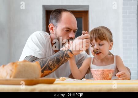 Père nourrissant sa fille avec une cuillère.Barbe chauve tatouée caucasien le père donne de la nourriture alimente son enfant fille petite fille blonde douce fille avec Banque D'Images