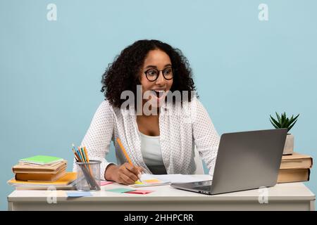 Concept d'enseignement à distance.Jeune femme noire excitée assise au bureau, utilisant un ordinateur portable, prenant des notes pendant le cours en ligne Banque D'Images