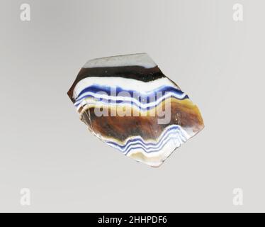 Fragment de bol nervuré en mosaïque de verre fin du 1st siècle C.-B.–début du 1st siècle A.D. fragment romain, probablement italien Rim.marron miel translucide, bleu cobalt et blanc opaque.rebord en saillie avec bord épais et arrondi;Côté droit en train de s'incliner vers le bas.motif en mosaïque de ruban formé de sections d'une canne simple au sol brun avec des fils irréguliers ondulés blancs et bleus dans des lignes horizontales parallèles; à l'extérieur, trois nervures verticales, espacées de manière inégale, avec des dessus aplatis et des bords extérieurs arrondis.intérieur poli; piqûres de bulles de surface et de fissures à l'intérieur;altération de la couche de mousse et d'irisée crémeuse o Banque D'Images