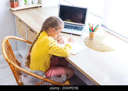 Un enfant écrit dans un carnet, assis au bureau près de la fenêtre.Une fille d'école blanche en sweat-shirt jaune fait ses devoirs à la maison devant un ordinateur portable.Accueil Banque D'Images