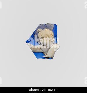 Fragment de caméo de verre fin du 1st siècle C.-B.–mi-1st siècle A.D. Roman bleu cobalt translucide avec revêtement blanc opaque.fragment de corps d'une tasse ou d'un bol relativement droit.en relief en blanc, tête d'une figure masculine jeune, peut-être Cupid, face à gauche mais regardant vers le bas,Avec le bras droit bien tendu avant lui et le bras gauche étendu vers le bas sur sa poitrine; un manteau fin, presque translucide, survole derrière sa tête.cassé de tous les côtés avec des fissures et des éclats à l'intérieur; mordillements, piqûres légères, et un léger vieillissement crémeux.marques de meulage rotatif à l'intérieur.Fragment de caméo en verre 249610 Banque D'Images