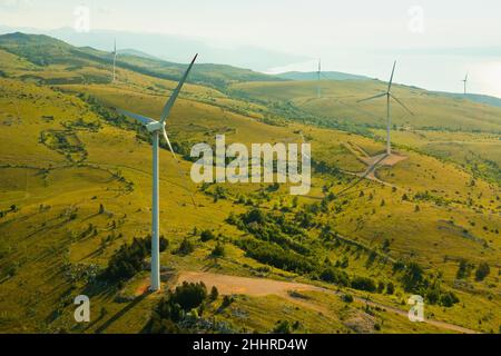Les moulins à vent fonctionnent à la station électrique sur les pentes de colline de haute montagne avec des arbres rares produisant de l'énergie verte près de la mer Adriatique dans le temps ensoleillé vue aérienne Banque D'Images