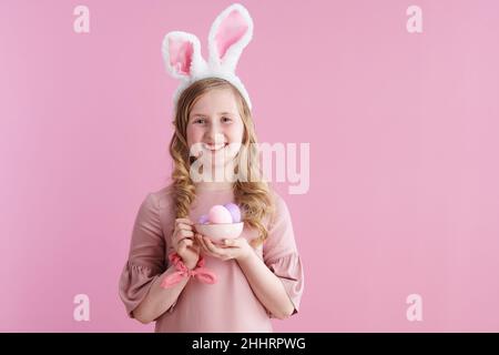 Portrait d'une jeune fille moderne souriante avec de longs cheveux blonds ondulés avec des oreilles de lapin et une tasse d'œufs de pâques isolés sur fond rose. Banque D'Images