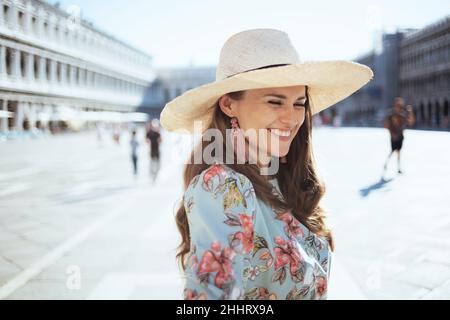 Femme élégante souriante en robe fleurie avec chapeau à la Piazza San Marco à Venise, Italie. Banque D'Images