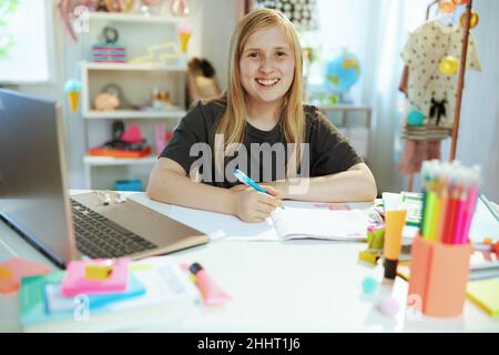 souriant moderne fille en chemise grise avec ordinateur portable et cahier d'exercices ayant l'éducation en ligne à la maison dans le soleil jour. Banque D'Images