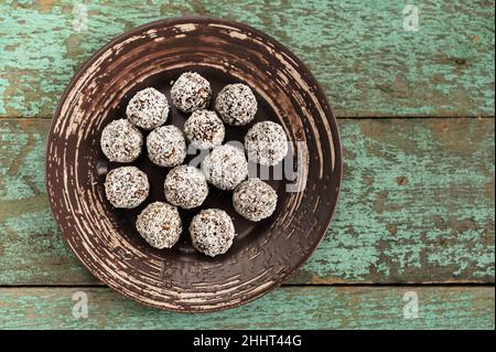 Bonbons végétaliens crus faits maison dans des copeaux de noix de coco dans une plaque en céramique sur une ancienne table en bois vue au plafond Banque D'Images