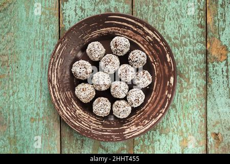 Bonbons végétaliens crus faits maison dans des copeaux de noix de coco dans une plaque en céramique sur une ancienne table en bois vue au plafond Banque D'Images