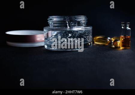Pot transparent avec gel hydratant cosmétique avec acide hyaluronique avec bulles et vitamines en capsules sur fond noir Banque D'Images