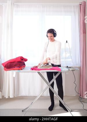 Femme repassant et écoutant de la musique avec le mobile dans une chambre Banque D'Images