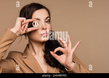 Magnifique femme montrant une pièce de monnaie dorée btc sur fond beige.Une femelle qui couvre l'œil avec un bitcoin, regarde l'appareil photo.Concept de blockchain de crypto-monnaie.Bourse, monnaies numériques monnaie et affaires Banque D'Images