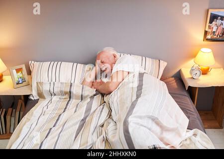 Homme âgé qui garde la tête sur l'oreiller tout en étant allongé sous une couverture rayée sur le lit double dans la chambre et dormant Banque D'Images