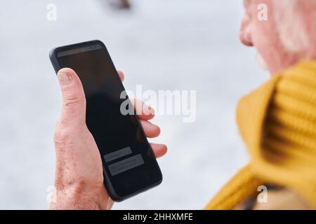 Main de l'homme âgé dans une écharpe tricotée jaune tenant un smartphone tout en lisant un nouveau message à l'extérieur le jour de l'hiver Banque D'Images