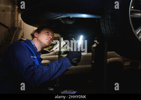 Gros plan d'un mécanicien, d'un technicien, d'un ingénieur automobile vérifie le soubassement de la voiture avec une lampe dans le garage d'un atelier de réparation.Réparation sous garantie du véhicule et Banque D'Images