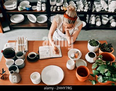 Le dur labeur ne s'arrête jamais.Photo en grand angle d'une femme méconnaissable assise et esquissant dans un carnet de son atelier de poterie. Banque D'Images